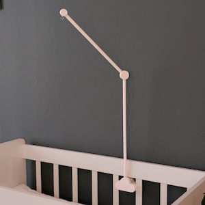 May include: A pale pink baby mobile arm is mounted on a dark gray wall above a matching crib. The mobile arm has a simple, minimalist design, with a curved top and a straight, adjustable arm.