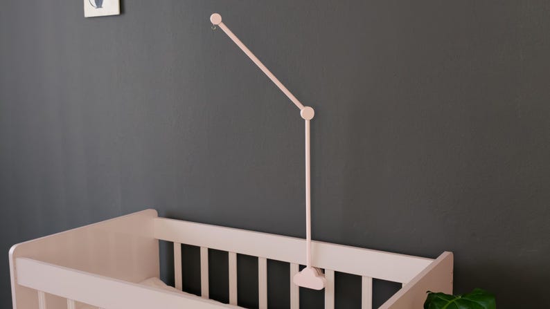 May include: A pale pink baby mobile arm is mounted on a dark gray wall above a matching crib. The mobile arm has a simple, minimalist design, with a curved top and a straight, adjustable arm.