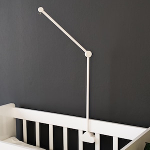 May include: A white baby crib with a white mobile arm attached to a dark gray wall. The mobile arm has a simple, modern design. A small framed picture hangs on the wall above the crib.