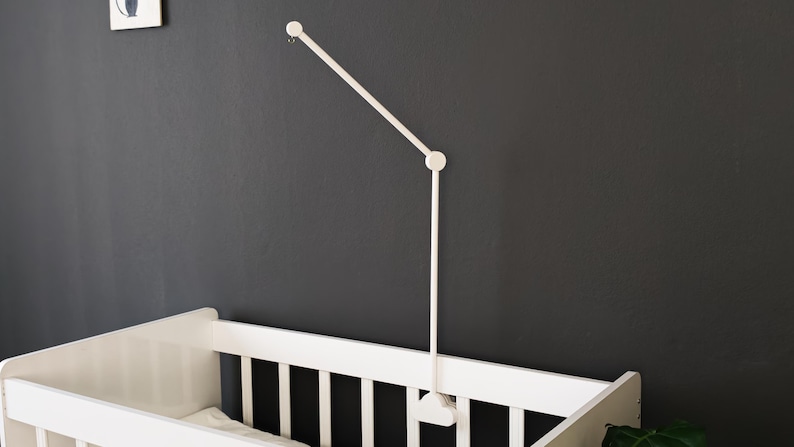 May include: A white baby crib with a white mobile arm attached to a dark gray wall. The mobile arm has a simple, modern design. A small framed picture hangs on the wall above the crib.