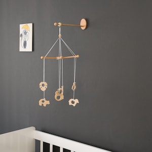 May include: Wooden baby mobile with hanging animal shapes, including a bear, elephant, and leaf. The mobile is suspended from a wooden arm attached to a dark grey wall. A cot is visible in the foreground.