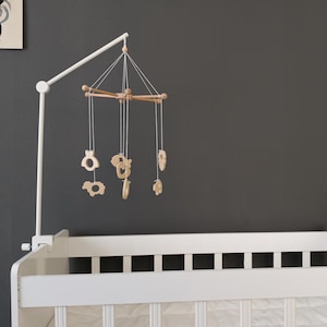May include: A white cot mobile arm attached to a white wooden cot, featuring a wooden cross-frame suspended by white strings. Several light-coloured wooden animal-shaped ornaments hang from the frame, creating a mobile that hangs above the cot.
