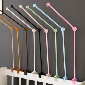 May include: A selection of baby mobile arms in a range of colours, including wood, brown, black, white, pink, green, and blue. Each arm is attached to a white cot and has a circular top. The arms are angled towards the viewer.