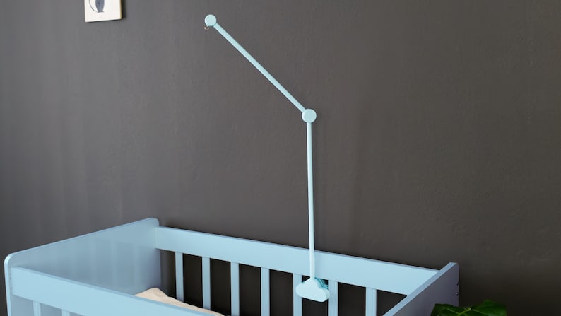 May include: A light blue baby crib with a matching mobile. The crib has a slatted design and is positioned against a dark gray wall. The mobile is attached to the wall and extends over the crib.
