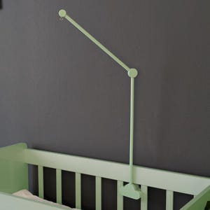 May include: A light green baby crib with a matching mobile arm attached to the wall. The crib has vertical slats and a simple design. The mobile arm is adjustable and extends over the crib. The wall is a dark gray color.