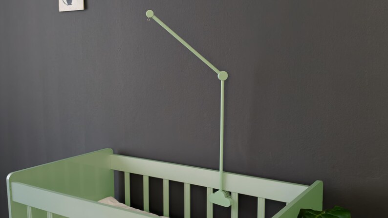 May include: A light green baby crib with a matching mobile arm attached to the wall. The crib has vertical slats and a simple design. The mobile arm is adjustable and extends over the crib. The wall is a dark gray color.