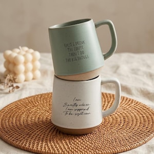 Puede incluir: Dos tazas de cerámica apiladas sobre una estera tejida. La taza superior es de color verde salvia con el texto "FIRST I DRINK THE COFFEE THEN I DO THE BIG THINGS." La taza inferior es blanca con el texto "I am Exactly where I am supposed to be right now."