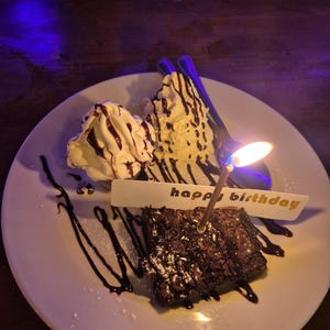 May include: A dessert plate with a brownie, vanilla ice cream, and chocolate syrup. A lit candle and a banner that says "happy birthday" are on the plate. The brownie is dusted with powdered sugar.