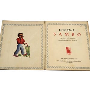 May include: Open book titled "Little Black Sambo" by Helen Bannerman, illustrated by Marjorie Romyns. The left page shows a cartoon figure in a red shirt, blue pants, and holding a green umbrella. The right page displays the title and publisher information.