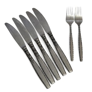 May include: A set of five silver-toned metal dinner knives and two matching forks. Each piece features a textured handle with a repeating pattern of circular swirls and small floral accents, paired with a smooth, plain metal blade or tines.