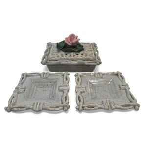May include: A ceramic set consisting of one lidded box and two square plates. The box features a sculpted pink rose with green leaves on the lid. All pieces have a light gray speckled finish and a decorative border resembling woven branches.