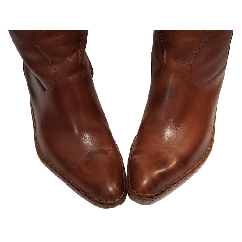 K&ouml;nnte beinhalten: Ein Paar braune Lederstiefel mit spitzer Zehenpartie. Die Stiefel haben eine mittlere Wadenh&ouml;he und einen leichten Absatz. Das Leder hat eine polierte Oberfl&auml;che und sichtbare N&auml;hte entlang der Sohlen. Die Stiefel sind vor wei&szlig;em Hintergrund.