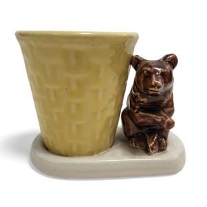 May include: A ceramic planter featuring a light yellow, basket-weave textured cup attached to a white oval base. A glossy brown bear figurine is positioned on the base, leaning against the side of the cup. The piece is made of glazed ceramic.