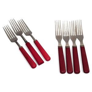 May include: A set of seven stainless steel forks featuring solid red handles. Three forks are arranged diagonally on the left, while four forks are positioned vertically on the right. Each fork has four tines and a smooth, cylindrical handle.