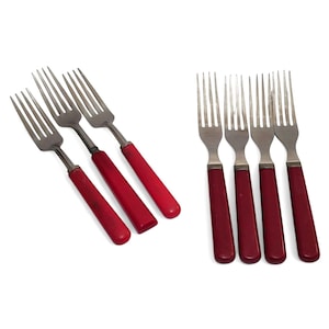 May include: A set of seven stainless steel forks featuring solid red handles. Three forks are arranged diagonally on the left, while four forks are positioned vertically on the right. Each fork has four tines and a smooth, cylindrical red handle.