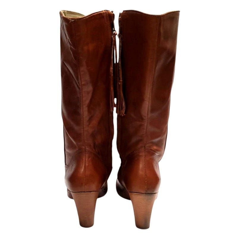 K&ouml;nnte beinhalten: Ein Paar braune kniehohe Lederstiefel. Die Stiefel haben einen Rei&szlig;verschluss mit Quastendetail und einen Holzabsatz. Die Stiefel sind aus glattem Leder gefertigt.