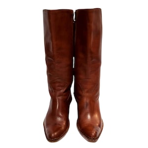 K&ouml;nnte beinhalten: Ein Paar hohe, braune Lederstiefel. Die Stiefel haben eine spitze Zehenpartie und einen Rei&szlig;verschluss an der Seite. Das Leder hat einen leicht abgenutzten Look. Die Stiefel sind kniehoch.