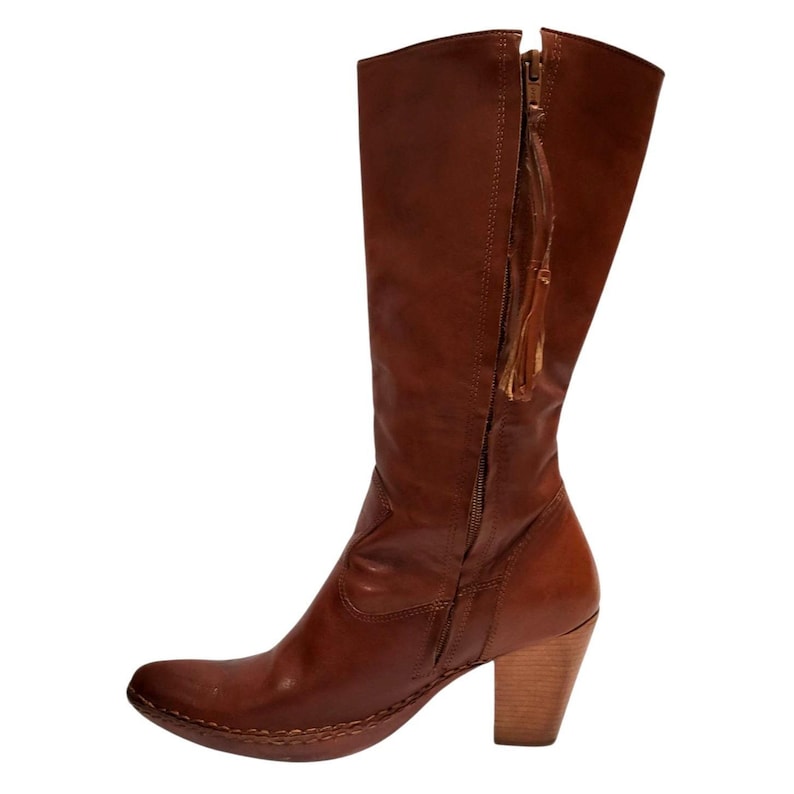 K&ouml;nnte beinhalten: Ein kniehoher Stiefel aus braunem Leder mit einem Holzabsatz. Der Stiefel hat einen seitlichen Rei&szlig;verschluss mit Quaste und eine runde Spitze. Der Absatz misst ca. 7,6 cm.