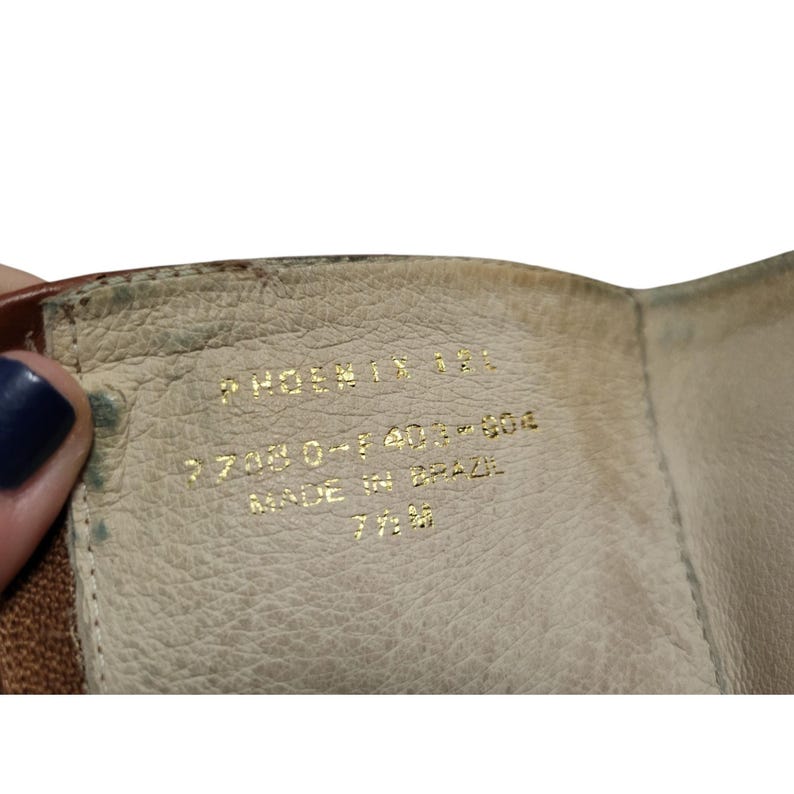 K&ouml;nnte beinhalten: Nahaufnahme des Inneren eines braunen Lederschuhs. Goldener Text lautet "PHOENIX 181", "773030-1401-804", "MADE IN BRAZIL" und "7 1/2 M". Das Innere des Schuhs ist hellbeige.