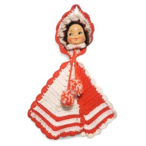 May include: A vintage handmade doll face kitchen towel holder. The doll has a painted face and is wearing a red and white crocheted hood and shawl. The shawl has red and white stripes and is trimmed with a scalloped edge. Two red and white pom-poms hang from the shawl.