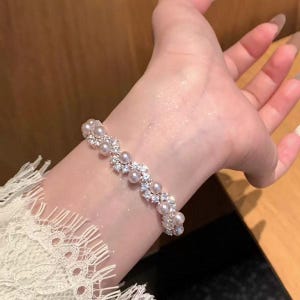 May include: A delicate bracelet featuring alternating clusters of small, round pearls and sparkling clear crystals. The bracelet is silver-toned and appears to be worn on a wrist. The background is a soft, warm tone.