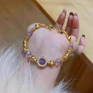 May include: Gold-coloured bracelet with a central round amethyst stone surrounded by small clear stones. The bracelet includes gold beads and purple crystal beads, with a wrapped gold wire design. The bracelet is held in a hand.