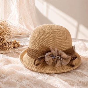 Op de afbeelding: Een beige hoed van gevlochten stro met een bruine mesh lintband rond de kroon. De hoed is versierd met een gelaagde stoffen bloem in een bijpassende bruine tint, die in het midden is voorzien van kleine ingelegde kristallen.