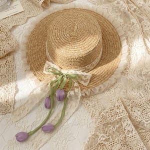 May include: A straw hat with a wide brim, adorned with a white lace ribbon and a green bow. The hat features a delicate lace trim and is embellished with three purple tulip accents, creating a charming and feminine accessory.