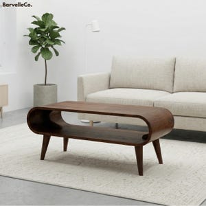 May include: A dark brown, mid-century modern coffee table with a unique, curved design. The table has a lower shelf and tapered legs. The table is in a living room setting with a light-colored sofa and a potted plant.