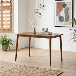 May include: A rectangular wooden dining table with four angled legs. The table is a warm brown color and is set in a well-lit room with a framed abstract art piece and a potted plant. The table is approximately 60 inches long.