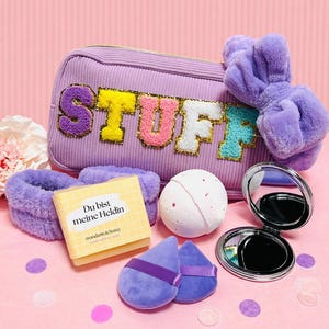 Könnte beinhalten: Eine Sammlung von Schönheits- und Pflegeprodukten, darunter eine lila Cord-Kosmetiktasche mit dem Wort "STUFF", ein flauschiges Stirnband, eine Badekugel, Puderquasten, ein Kompaktspiegel und ein Seifenstück mit deutschem Text.