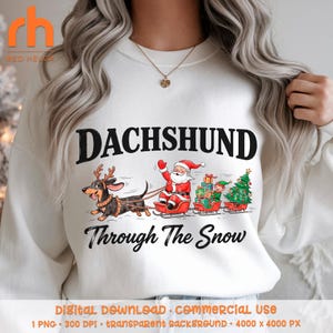 Puede incluir: Una sudadera blanca de cuello redondo con un gráfico de un perro salchicha con astas de reno tirando de un trineo rojo con Santa Claus, un elfo, regalos envueltos y un árbol de Navidad. El texto dice DACHSHUND Through The Snow.