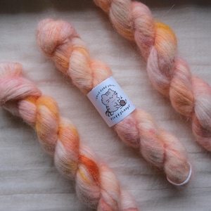 May include: Close-up of three skeins of hand-dyed yarn in shades of peach, orange, and yellow. Each skein has a label with a cartoon cat and the text "Fluffy Loops". The yarn appears soft and fluffy, ideal for knitting or crochet projects.