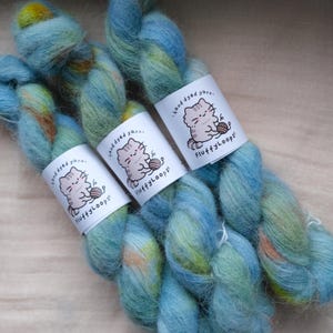 May include: Three skeins of hand-dyed yarn in a soft, fuzzy texture featuring a variegated color palette of sky blue, lime green, and hints of orange. Each skein is wrapped in a white paper label with a cartoon cat illustration and the text FluffyLoops.