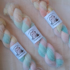 May include: Three skeins of hand-dyed yarn in a soft pastel palette of pink, yellow, and light blue. Each skein features a white paper label with the text 'hand dyed yarn' and 'FluffyLoops' alongside a graphic illustration of a cat holding yarn.