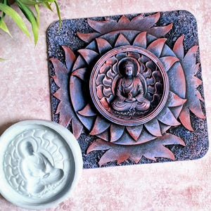 May include: A detailed, multi-layered wall art piece featuring a seated Buddha figure in a lotus position, surrounded by a floral design. The color palette includes shades of red, blue, and gray. A white, circular mold of the Buddha is also present.