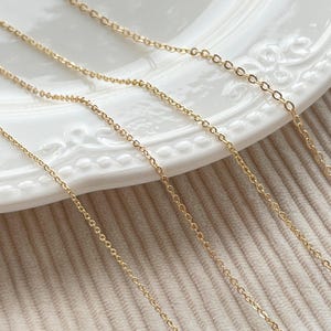 Può includere: Primo piano di diverse delicate collane a catena color oro. Le catene sono esposte su un piatto bianco decorativo e su una superficie beige strutturata. L'immagine include un logo blu e bianco che recita "Zentro DIY Supply".
