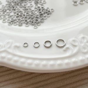 Op de afbeelding: Zilverkleurige sprongringen in verschillende maten, verspreid op een wit, decoratief bord. De afbeelding bevat een logo met de tekst "Zentro DIY Supply" en "EST 2023". Ideaal voor het maken van sieraden en knutselen.