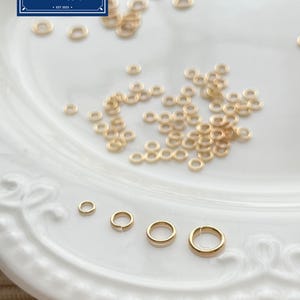 Puede incluir: Anillos de salto dorados de varios tamaños esparcidos sobre un plato blanco con borde. Los anillos son para la fabricación de joyas DIY. La imagen incluye el texto "Zentro DIY Supply EST 2023" en un recuadro azul y blanco.