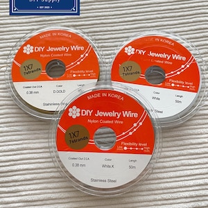 May include: Three circular spools of DIY Jewelry Wire, nylon coated, 1x7 strands, 0.38 mm diameter, 50m length. Labels indicate colors D.GOLD, White, and White.K, with stainless steel core. Made in Korea, featuring flexibility level indicators.