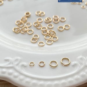 Può includere: Anelli di giunzione dorati di varie dimensioni su un piatto bianco. Questi anelli aperti sono adatti per la creazione di gioielli e altri progetti artigianali. L'immagine include un logo che dice "Zentro DIY Supply".