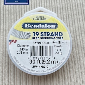 Op de afbeelding: Een ronde spoel Beadalon 19-strengs rijgdraad voor kralen in satijngoud. Het label vermeldt een diameter van 0,38 mm, een breeksterkte van 5 kg en een lengte van 9,2 m. De spoel bevat een streepjescode en de productcode JW14NG-0.