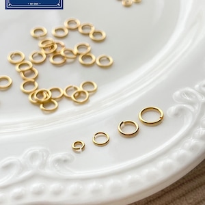 Puede incluir: Anillos de salto dorados de varios tamaños están dispersos sobre un plato blanco decorativo. Los anillos están dispuestos en grupos e individualmente, mostrando su forma circular. El logotipo de Zentro DIY Supply está en la esquina superior izquierda.