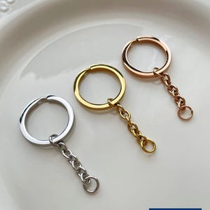 Op de afbeelding: Drie sleutelhangers in zilver, goud en roségoud. Elke sleutelhanger heeft een ronde ring en een korte ketting met een kleine ring aan het uiteinde. De sleutelhangers worden gepresenteerd op een wit bord. De rechter benedenhoek heeft een blauw en wit logo met de tekst "Zentro DIY Supply".