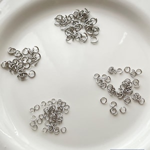 Puede incluir: Primer plano de un plato de cerámica blanca con borde decorativo, que contiene varias pilas de pequeños anillos de salto plateados. Los anillos están dispersos por el plato, listos para proyectos de bricolaje. Un logotipo azul y blanco está en la esquina inferior derecha.