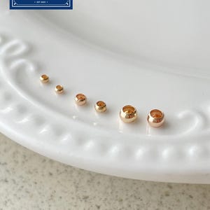 Könnte beinhalten: Sechs kleine, runde, metallisch goldfarbene Perlen, die der Größe nach in einer Reihe angeordnet sind. Die Perlen haben ein Loch in der Mitte und eine glatte, reflektierende Oberfläche. Ein blaues Logo oben links zeigt den Text Zentro DIY Supply.