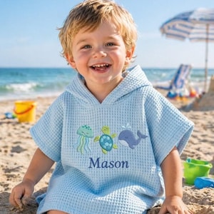 May include: A light blue waffle-weave hooded beach towel with embroidered sea creatures and the name "Mason". The towel features a jellyfish, turtle, and whale design. The child is smiling on a sandy beach.