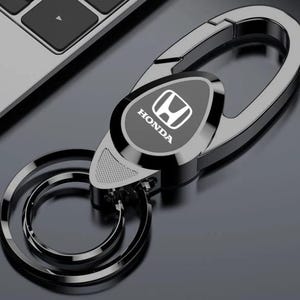 Puede incluir: Un llavero Honda gris metálico con un emblema ovalado negro con el logotipo de Honda en blanco. El llavero tiene un cierre grande en forma ovalada y dos anillos más pequeños. El diseño es elegante y moderno.