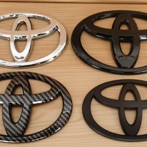 Op de afbeelding: Vier Toyota-emblemen in verschillende stijlen. De linkerbovenkant is chroom, de rechterbovenkant is glanzend zwart, de linksonderkant is koolstofvezel en de rechteronderkant is matzwart. Allemaal ovaal met het Toyota-logo.