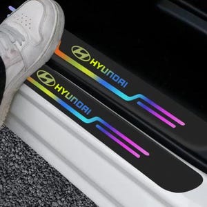 May include: Black car door sill protectors with the Hyundai logo and text. The logo and text are illuminated with a gradient of colors. A person's foot is visible stepping onto the sill.